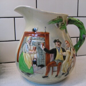 May include: Cream-colored ceramic pitcher with a green, vine-like handle. The pitcher features a relief scene of people in a pub, with figures in period clothing. The handle is a vibrant green, and the spout is curved.