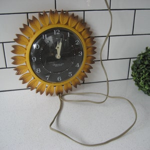 Op de afbeelding: Een gele zonnebloemvormige klok met zwarte cijfers en een zwart gezicht. De klok heeft een snoer en is aangesloten. De klok staat op een wit betegeld oppervlak.