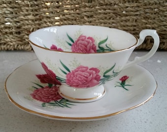 May include: A white teacup and saucer set with a pink carnation floral pattern. The cup and saucer have a gold trim around the edge.