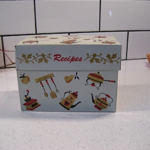 May include: White metal recipe box with a red and gold design. The box has a lid with the word "Recipes" printed in red. The box is decorated with images of kitchen utensils, teapots, and other kitchen items.