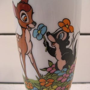 May include: A white ceramic mug featuring a cartoon image of Bambi and Flower from Disney's Bambi. Bambi is a brown deer with large ears and brown eyes. Flower is a skunk with black and white fur and a pink nose. They are both holding flowers. The mug has the text "Walt Disney Productions" at the bottom.