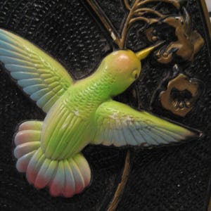 K&ouml;nnte beinhalten: Ein gr&uuml;ner, blauer und rosa Kolibri mit ausgebreiteten Fl&uuml;geln ist auf schwarzem Hintergrund dargestellt. Der Kolibri sitzt auf einem goldenen Zweig mit goldenen Bl&uuml;ten.