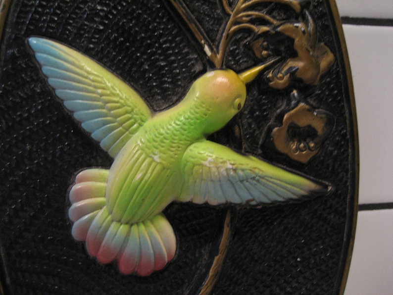 K&ouml;nnte beinhalten: Ein gr&uuml;ner, blauer und rosa Kolibri mit ausgebreiteten Fl&uuml;geln ist auf schwarzem Hintergrund dargestellt. Der Kolibri sitzt auf einem goldenen Zweig mit goldenen Bl&uuml;ten.