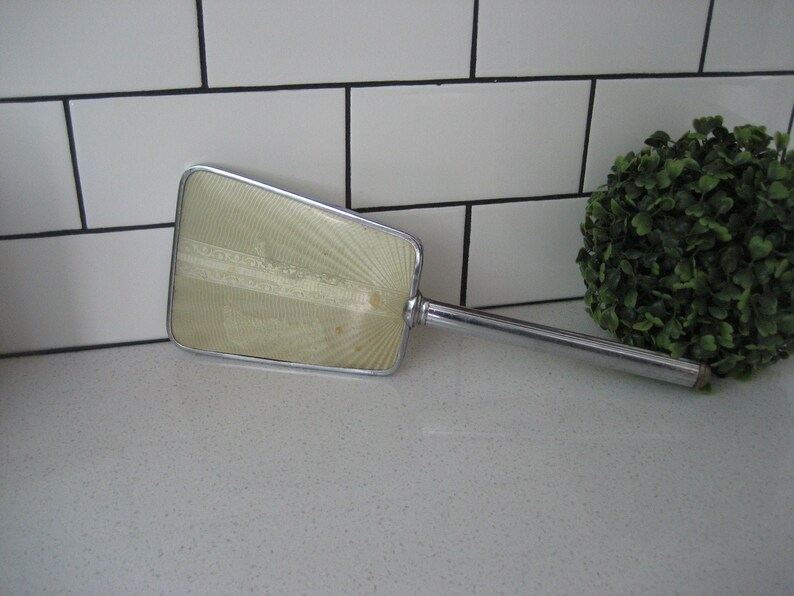 May include: A vintage hand mirror with a silver handle and a clear, slightly yellowed, rectangular mirror with a decorative pattern. The mirror is lying on a white countertop.