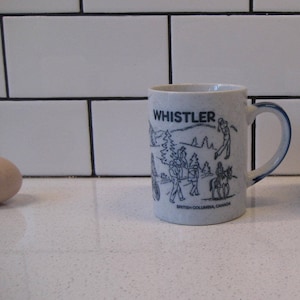 May include: White ceramic mug with a blue rim and handle. The mug features a black and white illustration of people skiing and snowboarding in the mountains. The text "WHISTLER" is printed in blue on the mug. The text "BRITISH COLUMBIA, CANADA" is printed in blue at the bottom of the mug.