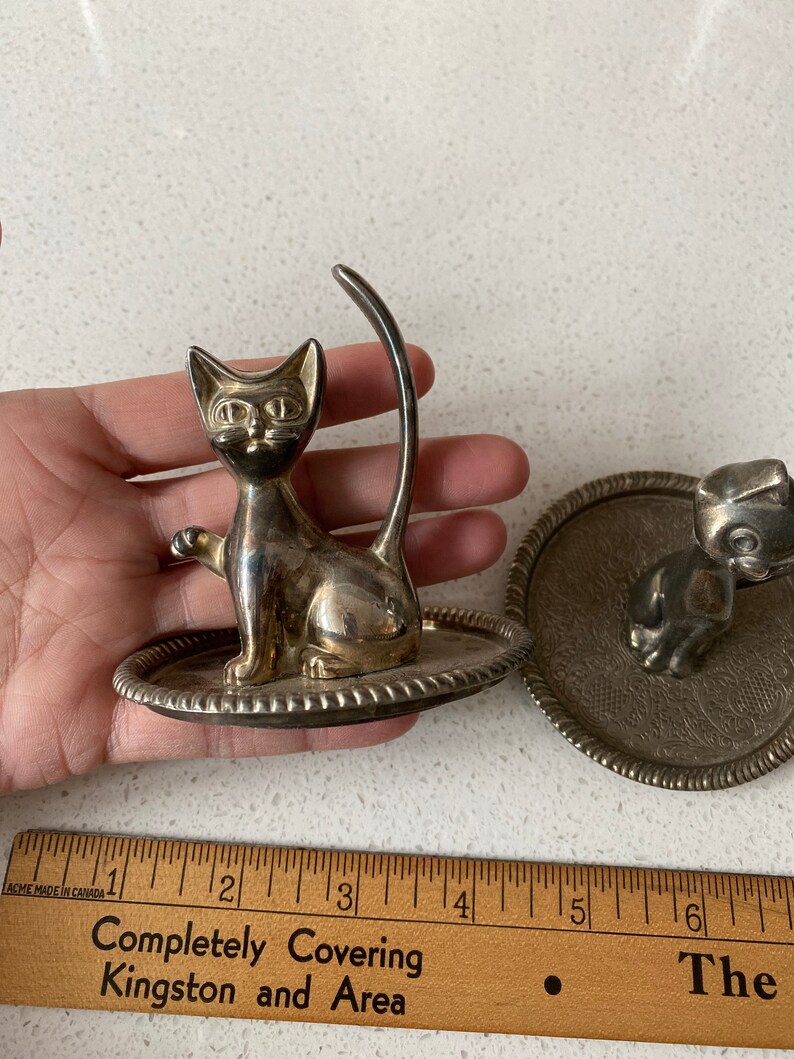 K&ouml;nnte beinhalten: Zwei silberne Ringhalter, einer in Form einer Katze und der andere in Form eines Hundes. Beide Halter haben eine kleine Schale, um den Ring zu halten.