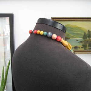 May include: A colorful beaded necklace with wooden beads and a variety of colors, including yellow, green, blue, and pink. The necklace is on a dark gray mannequin.