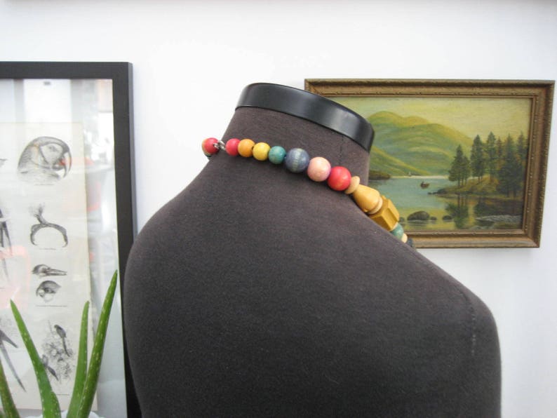 May include: A colorful beaded necklace with wooden beads and a variety of colors, including yellow, green, blue, and pink. The necklace is on a dark gray mannequin.