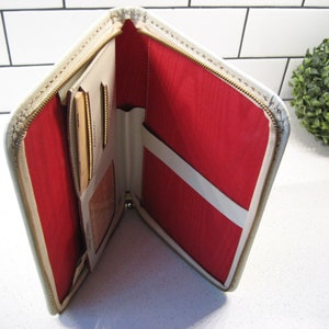 May include: A white leather zippered travel wallet with a red interior. The wallet has multiple compartments for cards and cash.