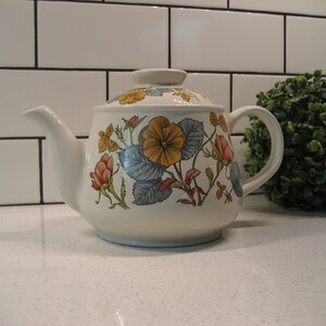 May include: A white ceramic teapot with a floral design featuring yellow, orange, and pink flowers on a blue and green leafy background.