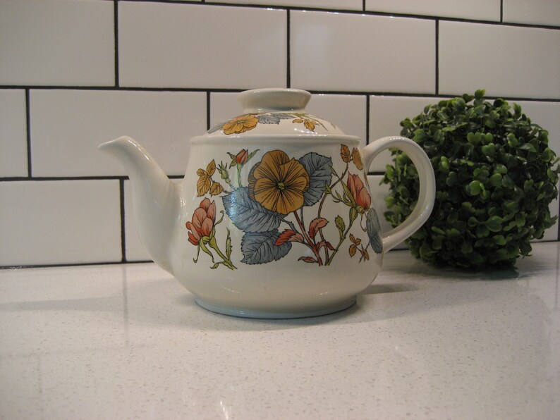 May include: A white ceramic teapot with a floral design featuring yellow, orange, and pink flowers on a blue and green leafy background.