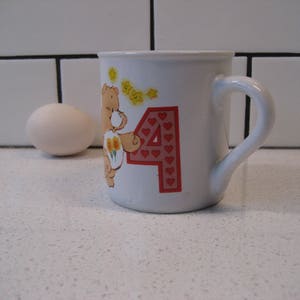 May include: White ceramic mug with a cartoon bear holding flowers and the number 4 with red hearts. The bear is brown with yellow flowers.
