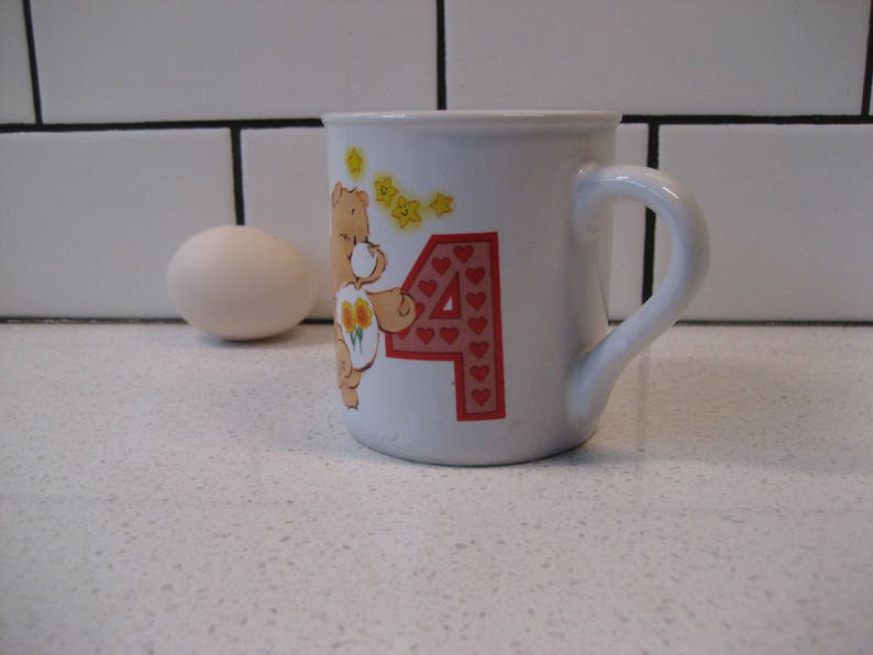May include: White ceramic mug with a cartoon bear holding flowers and the number 4 with red hearts. The bear is brown with yellow flowers.