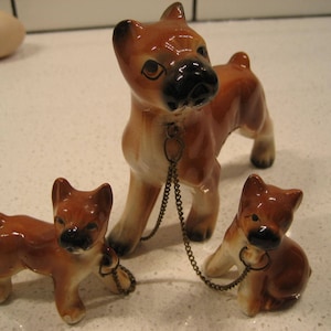 May include: Three ceramic figurines of boxer dogs. The largest dog is brown and black with a chain around its neck. The two smaller dogs are brown and black and are standing on either side of the larger dog.