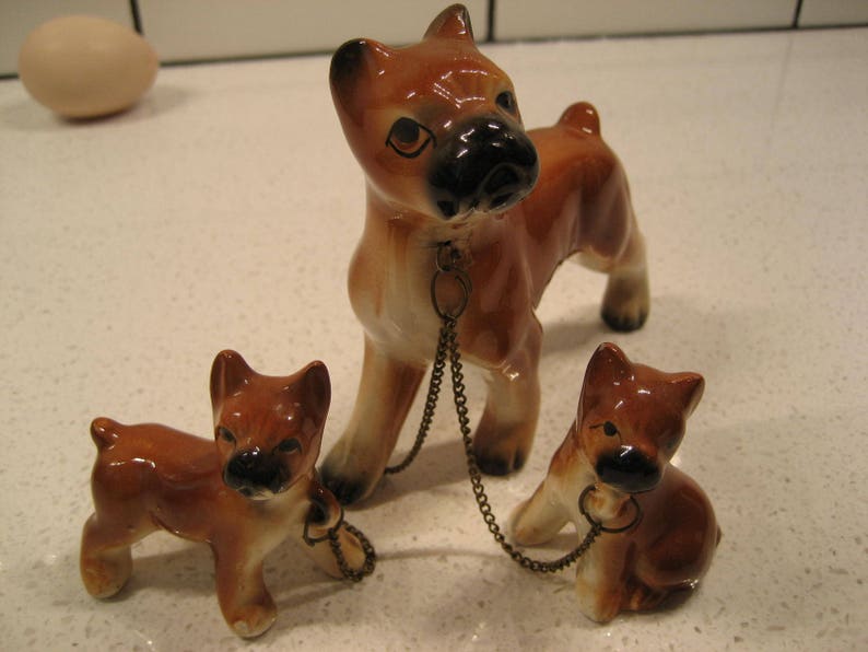 May include: Three ceramic figurines of boxer dogs. The largest dog is brown and black with a chain around its neck. The two smaller dogs are brown and black and are standing on either side of the larger dog.