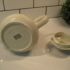 May include: White ceramic teapot with a lid and a handle. The bottom of the teapot has the words "Sadler England" printed on it.