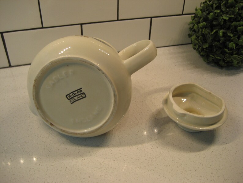 May include: White ceramic teapot with a lid and a handle. The bottom of the teapot has the words "Sadler England" printed on it.