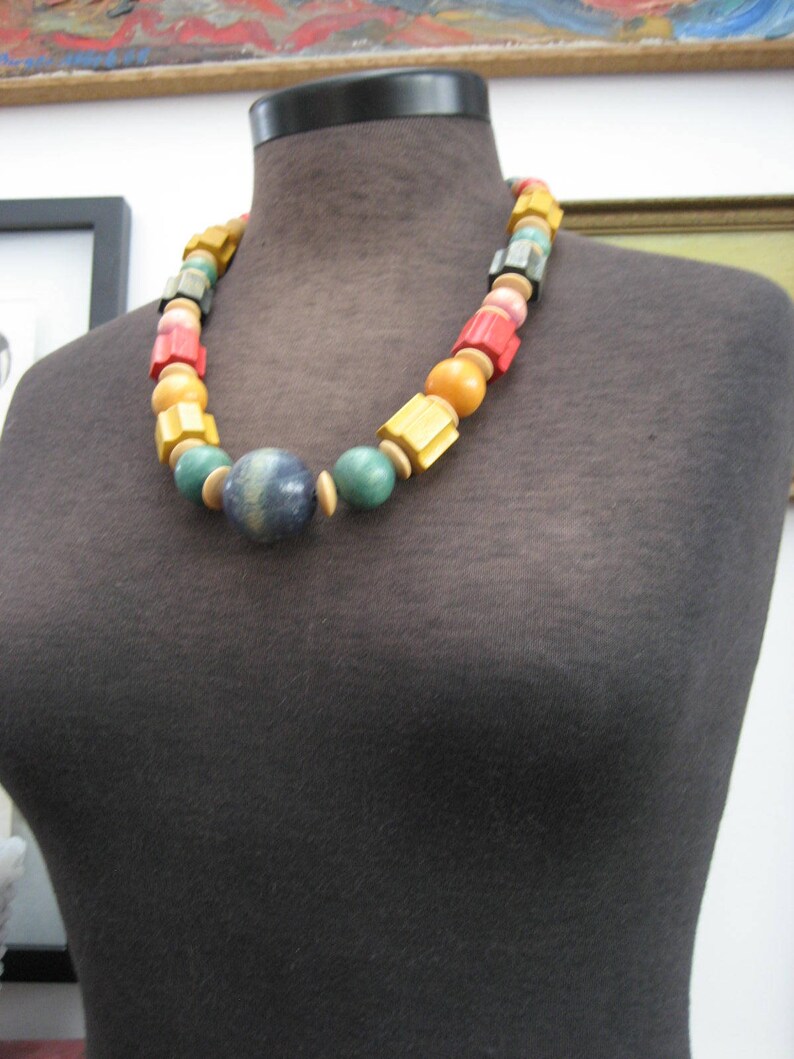 May include: A colorful wooden bead necklace with a blue and green bead in the center. The necklace is on a black mannequin.