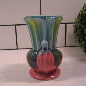 May include: A small, colourful ceramic vase with a pink base, green and blue stripes, and a pink flower design.