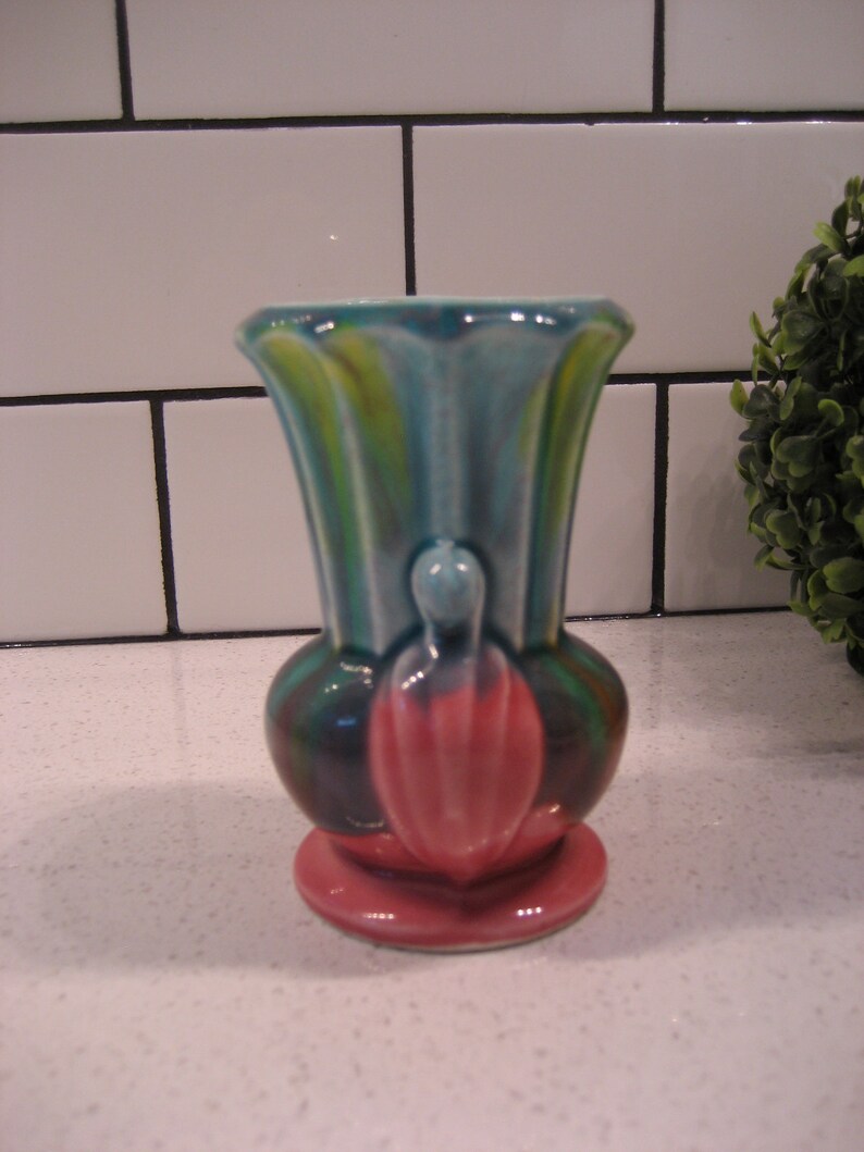 May include: A small, colourful ceramic vase with a pink base, green and blue stripes, and a pink flower design.