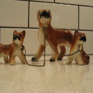 May include: Three ceramic figurines of brown and white boxer dogs. The largest dog is standing and has a gold chain attached to its collar. The two smaller dogs are sitting and are connected to the chain by a ring on their collars.