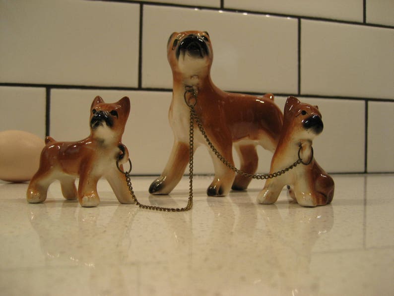 May include: Three ceramic figurines of brown and white boxer dogs. The largest dog is standing and has a gold chain attached to its collar. The two smaller dogs are sitting and are connected to the chain by a ring on their collars.