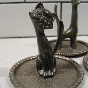 K&ouml;nnte beinhalten: Zwei silberne Ringhalter, einer in Form einer Katze und der andere in Form einer Giraffe. Beide stehen auf kleinen, runden, silbernen Tabletts mit einem verzierten Design.