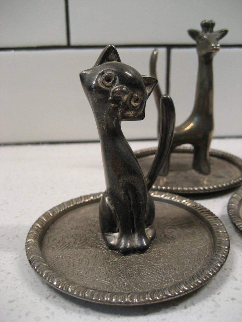 K&ouml;nnte beinhalten: Zwei silberne Ringhalter, einer in Form einer Katze und der andere in Form einer Giraffe. Beide stehen auf kleinen, runden, silbernen Tabletts mit einem verzierten Design.