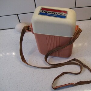 Peut inclure: Une boîte à lunch rose et blanche avec une sangle marron et blanche. La boîte à lunch porte une étiquette indiquant "Thermostyle Lunch Pack".