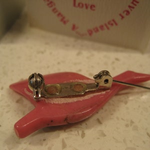 May include: A pink plastic brooch shaped like a bird with a silver metal clasp and pin. The brooch has a white stripe across the body.