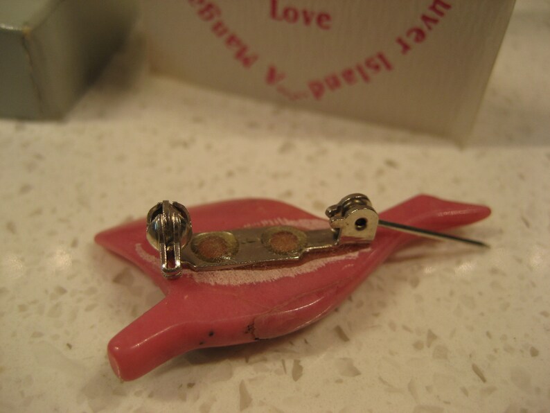 May include: A pink plastic brooch shaped like a bird with a silver metal clasp and pin. The brooch has a white stripe across the body.