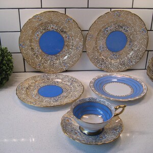 May include: A vintage china set featuring gold filigree, blue details, and gold rims. The set includes plates, saucers, and a teacup. The plates have a blue centre. The teacup and saucer have gold handles and rims.