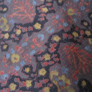May include: A close-up of a fabric with a repeating pattern of black, red, blue, and yellow floral designs. The fabric has a textured, woven appearance.