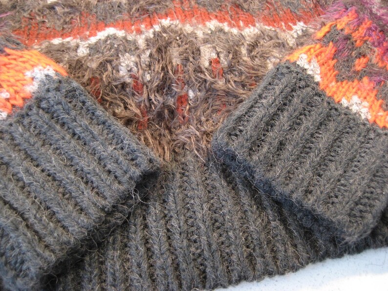 May include: A close-up of a gray knitted sweater with orange and white stripes. The sweater has a fuzzy texture and is made of a thick yarn.