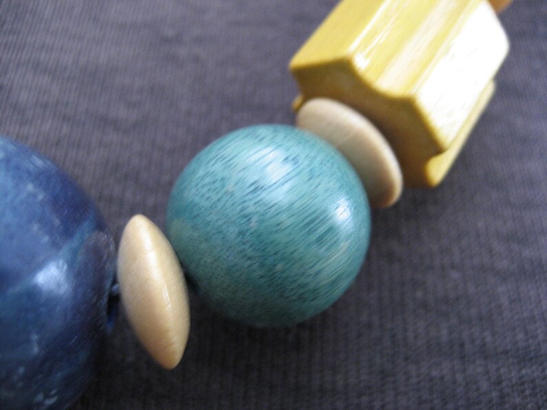 May include: A close-up of a necklace made of wooden beads. The beads are blue, green, and cream colored. The beads are round and have a smooth finish.