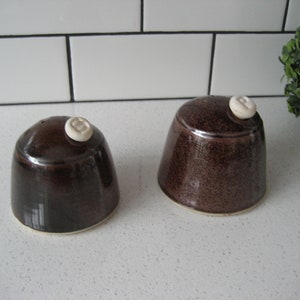 Puede incluir: Dos saleros y pimenteros de cerámica marrón con tapas de cerámica blanca. Los saleros tienen un esmalte moteado y una forma redondeada.