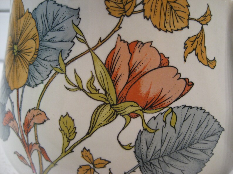 May include: A vintage ceramic pitcher with a floral design featuring a large pink rose, yellow flowers, and green leaves on a white background.