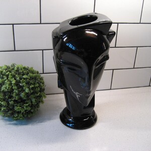 May include: A black ceramic vase shaped like a stylised human head. The vase has a smooth, glossy finish and a wide opening at the top.