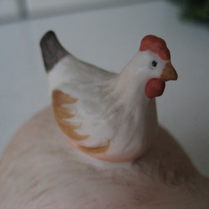 May include: A ceramic hen figurine with a white body, brown wing accents, and a red comb and wattle. The hen is perched atop a larger, light pink form, possibly a decorative piece or part of a set. The figurine has a detailed, hand-painted appearance.