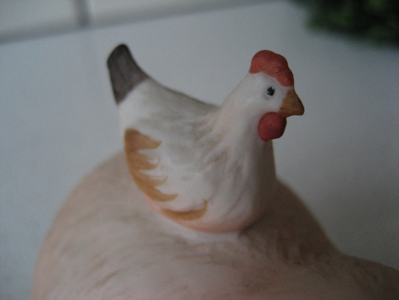May include: A ceramic hen figurine with a white body, brown wing accents, and a red comb and wattle. The hen is perched atop a larger, light pink form, possibly a decorative piece or part of a set. The figurine has a detailed, hand-painted appearance.