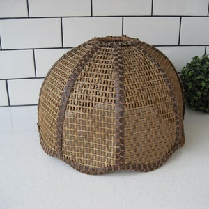 May include: A woven lampshade with a rounded, dome-like shape. The shade is made of light brown wicker with darker brown trim and accents. The design features a repeating square pattern.