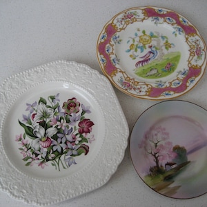 May include: Three decorative ceramic plates with floral and nature-inspired designs. The largest plate features a white background with a pink, purple, and green floral design. The medium-sized plate has a pink and gold border with a white background and a yellow and green bird design. The smallest plate has a pink and white background with a pink and green tree design.