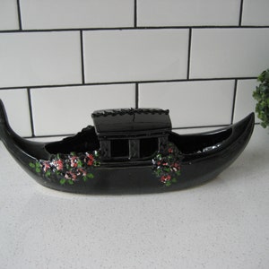 May include: Black ceramic gondola planter with a painted floral design. The planter has a small cabin-like structure on the top.