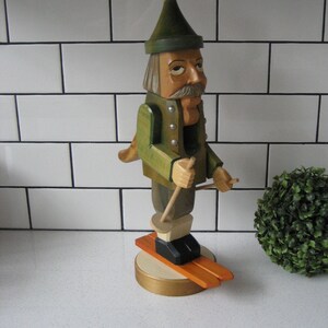 Könnte beinhalten: Eine Holzfigur eines Skifahrers mit grünem Hut und Jacke, der Skistöcke hält. Die Figur steht auf einem Holzsockel mit orangenen Skiern.