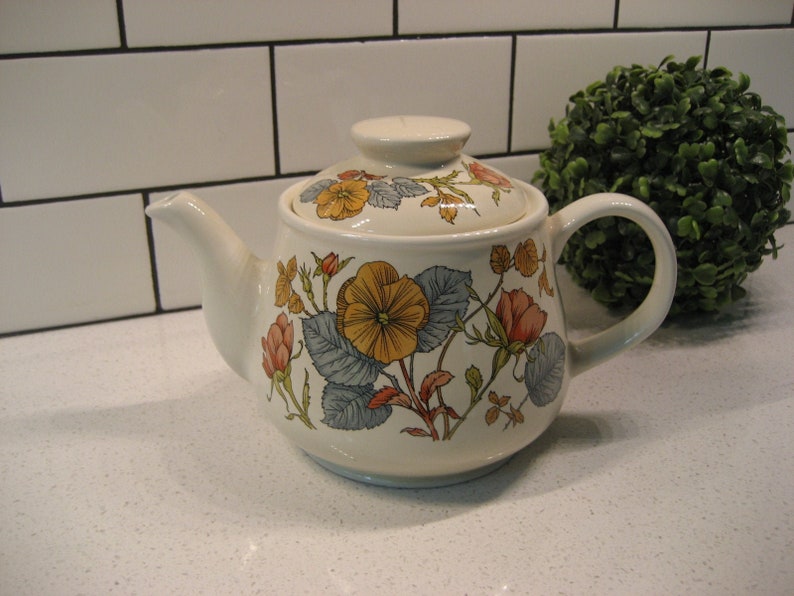 May include: A white ceramic teapot with a floral design featuring yellow, orange, and pink flowers. The teapot has a lid and a handle.