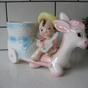 May include: A vintage ceramic planter shaped like a donkey with a little boy riding on its back. The boy is wearing a yellow hat and holding a blue and white toy. The planter is painted in shades of pink, blue, and white.