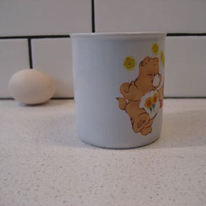 May include: White ceramic mug with a cartoon illustration of a brown bear wearing a yellow flower crown and holding a bouquet of yellow flowers. The bear is surrounded by yellow stars. The mug has a white interior and a rounded handle.
