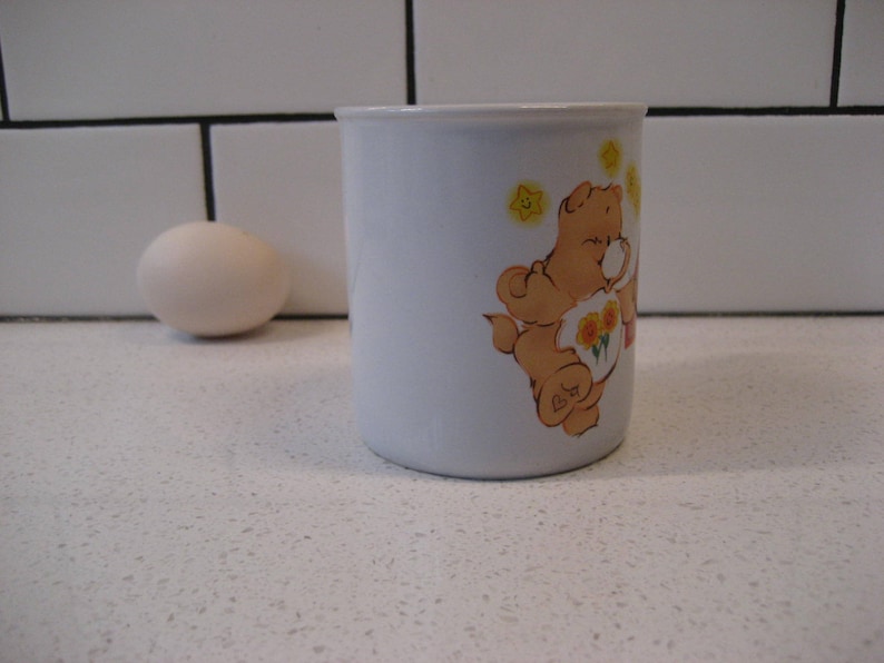 May include: White ceramic mug with a cartoon illustration of a brown bear wearing a yellow flower crown and holding a bouquet of yellow flowers. The bear is surrounded by yellow stars. The mug has a white interior and a rounded handle.