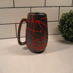 K&ouml;nnte beinhalten: Eine schwarze Keramiktasse mit einem roten abstrakten Design. Die Tasse hat einen Henkel und ist etwa 15 cm hoch.