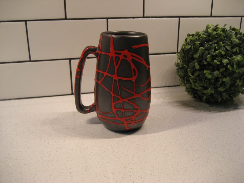 K&ouml;nnte beinhalten: Eine schwarze Keramiktasse mit einem roten abstrakten Design. Die Tasse hat einen Henkel und ist etwa 15 cm hoch.